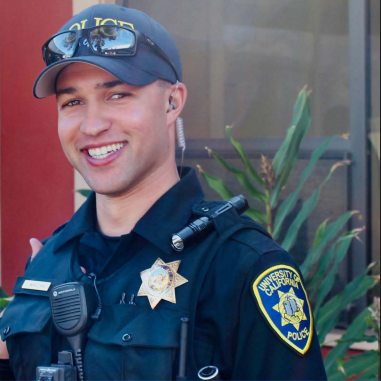 A smiling university police officer in uniform stands outdoors, wearing a cap, radio, and sunglasses on his hat.
